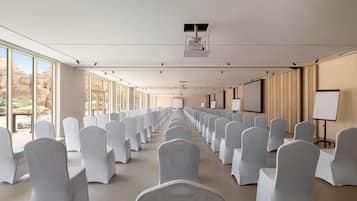 Un salón de conferencias con filas de sillas blancas, ventanas amplias y un proyector en el techo.