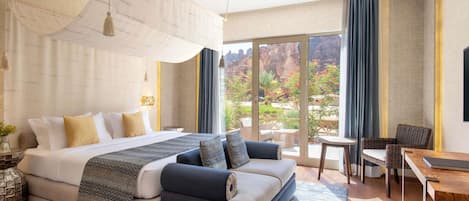 A modern hotel room with a large bed, a sofa, a dining area, and a view of a mountainous landscape.