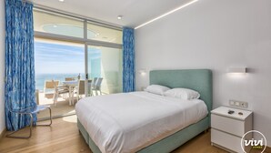A modern bedroom with a large bed, a nightstand, and a view of the ocean through floor-to-ceiling windows. - Nova | Panoramic Sea View - Pool (Fuengirola)