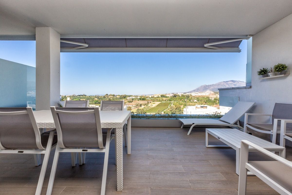 A balcony with a table and chairs, a view of a landscape with buildings and a mountain.