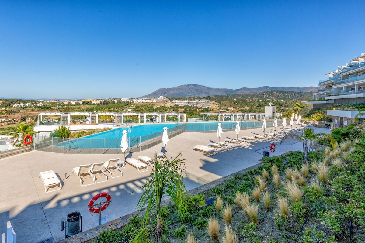 A rooftop pool area with lounge chairs and umbrellas, surrounded by greenery and overlooking a mountainous landscape.