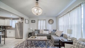 A modern living room with a sofa, coffee table, and a chandelier. There is a kitchen area with cabinets and a refrigerator.