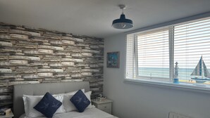 Une chambre avec un mur en pierre, un lit recouvert d’une couvre-lit à motifs, une table de chevet, une lampe et une fenêtre donnant sur la mer.