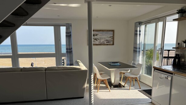 Un salon moderne comprenant un canapé, une table à manger et des chaises, un coin cuisine et une grande fenêtre donnant sur une plage.