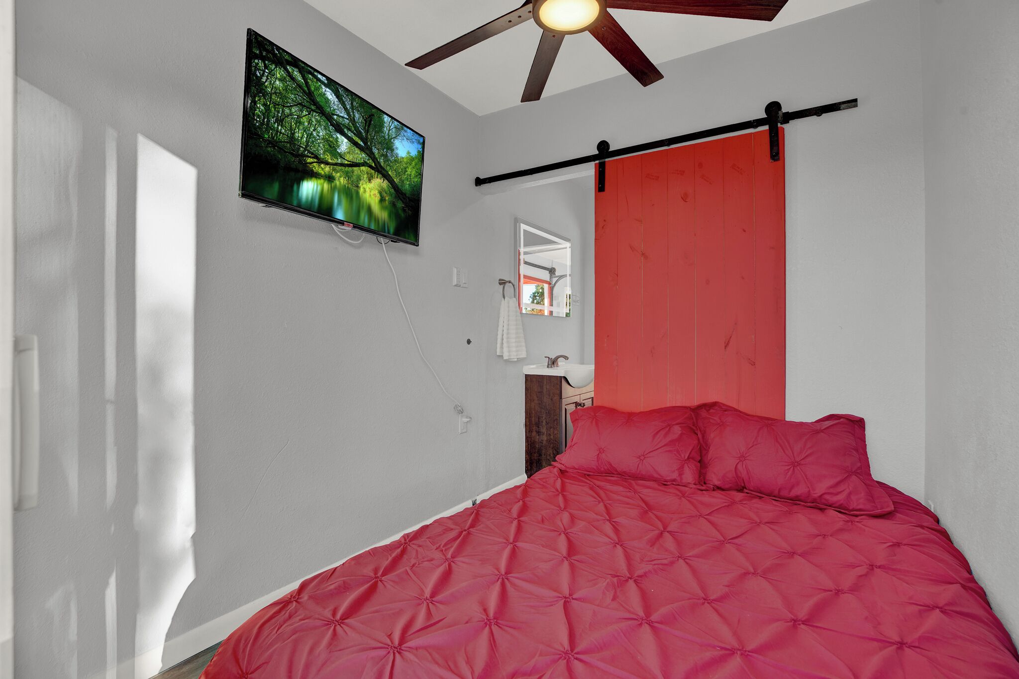 A bedroom with a red bedspread, a ceiling fan, a TV mounted on the wall, and a red sliding door.