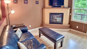 A living room with a leather sofa, a coffee table, a fireplace, a television, and a large window. - Abbey Chateau Treehouse #10 / Oak Crest (Eureka Springs)