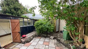 A paved patio with a wooden door, a small garden area with a bush, and a green trash bin.