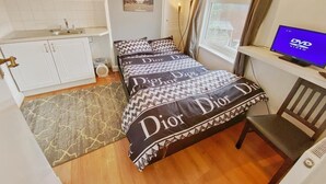 A bedroom with a bed, a small desk with a computer, a window with curtains, a sink, and a toilet.