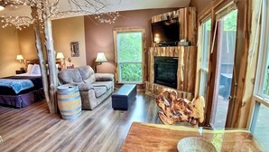 Une pièce confortable, aménagée comme un chalet, avec un plancher en bois, un canapé, une petite table, un téléviseur fixé à un mur en bois et une grande fenêtre donnant sur l’extérieur.