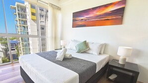 Una habitación de hotel moderna con una cama grande, dos mesitas de noche con lámparas, un cuadro de un atardecer y vistas de un paisaje urbano a través de amplias ventanas.