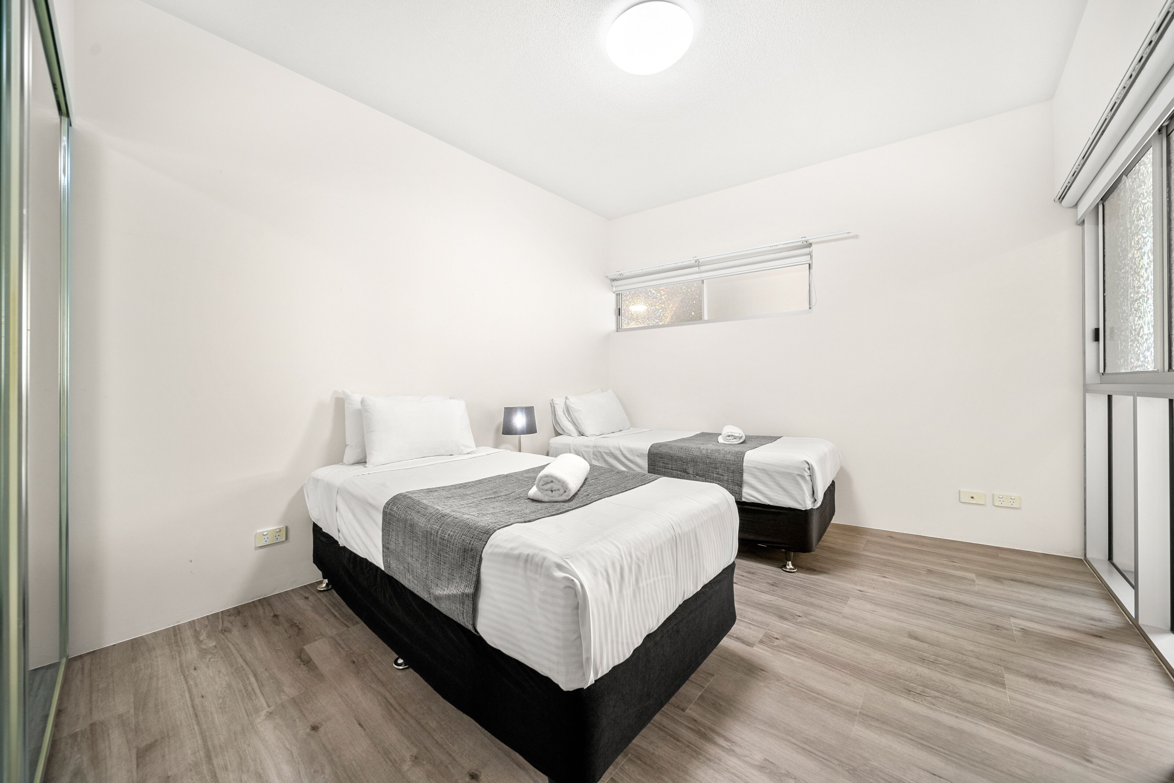 A hotel room with two beds, a small bedside table with a lamp, a mirror above the beds, and a window with blinds.
