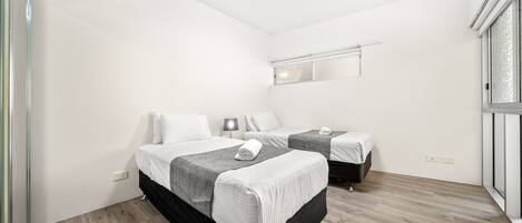 A hotel room with two beds, a small bedside table with a lamp, a mirror above the beds, and a window with blinds.