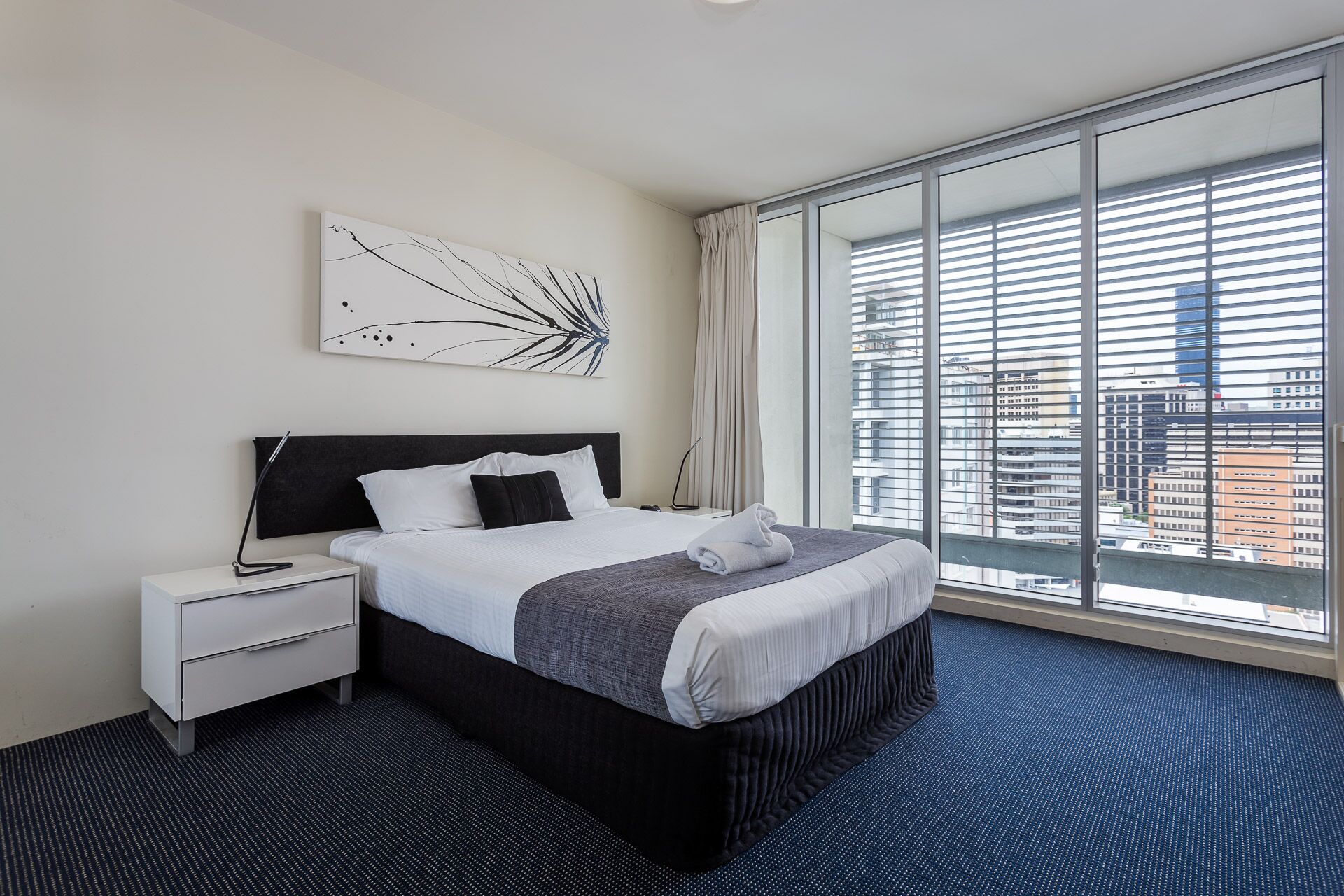 Camera d'hotel con un letto grande, un comodino, un'opera d'arte moderna e vista sulla città attraverso ampie finestre a tutta parete.
