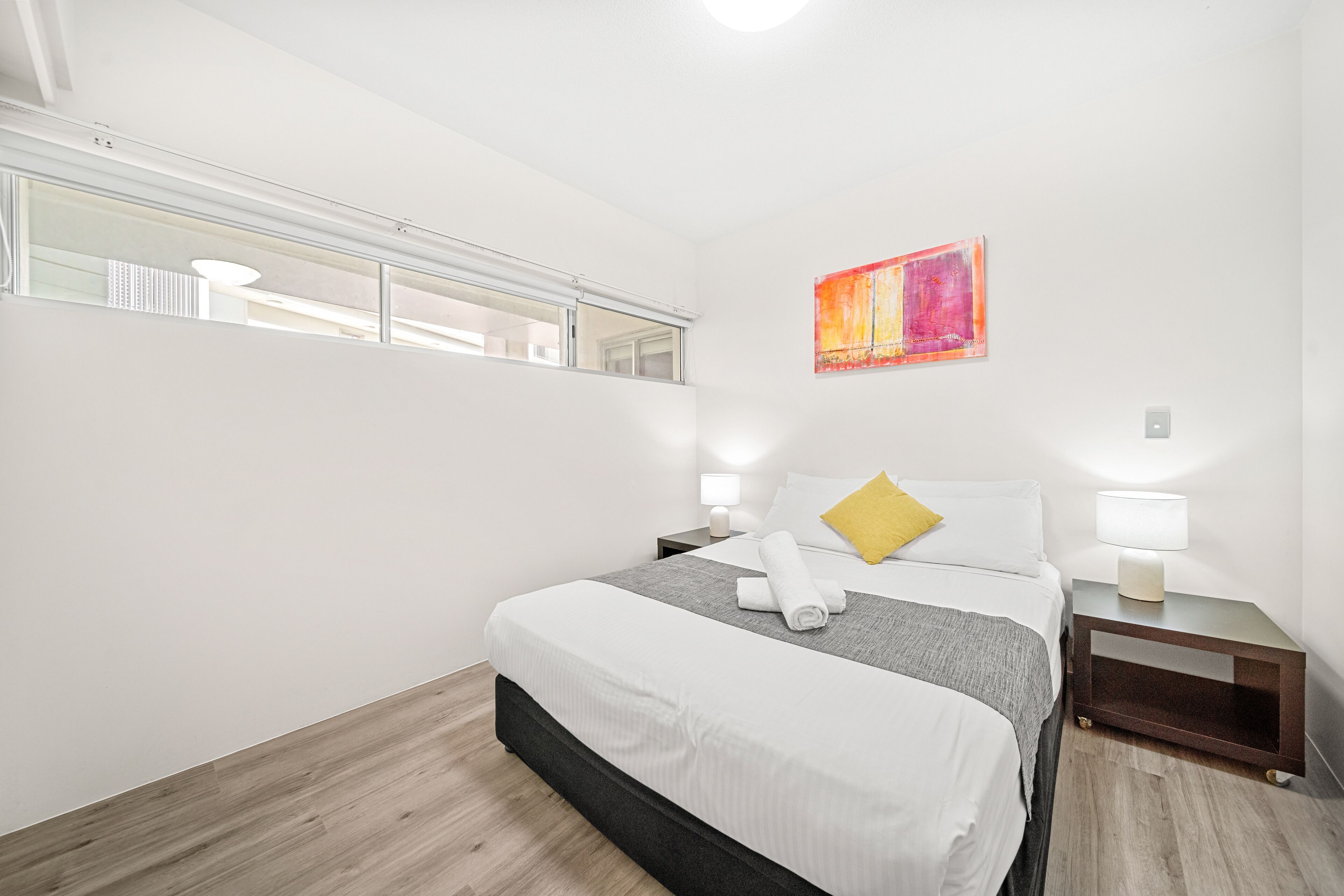 A modern hotel room with a large bed, a bedside table with a lamp, a small bedside table with a lamp, a window with blinds, and a painting on the wall.