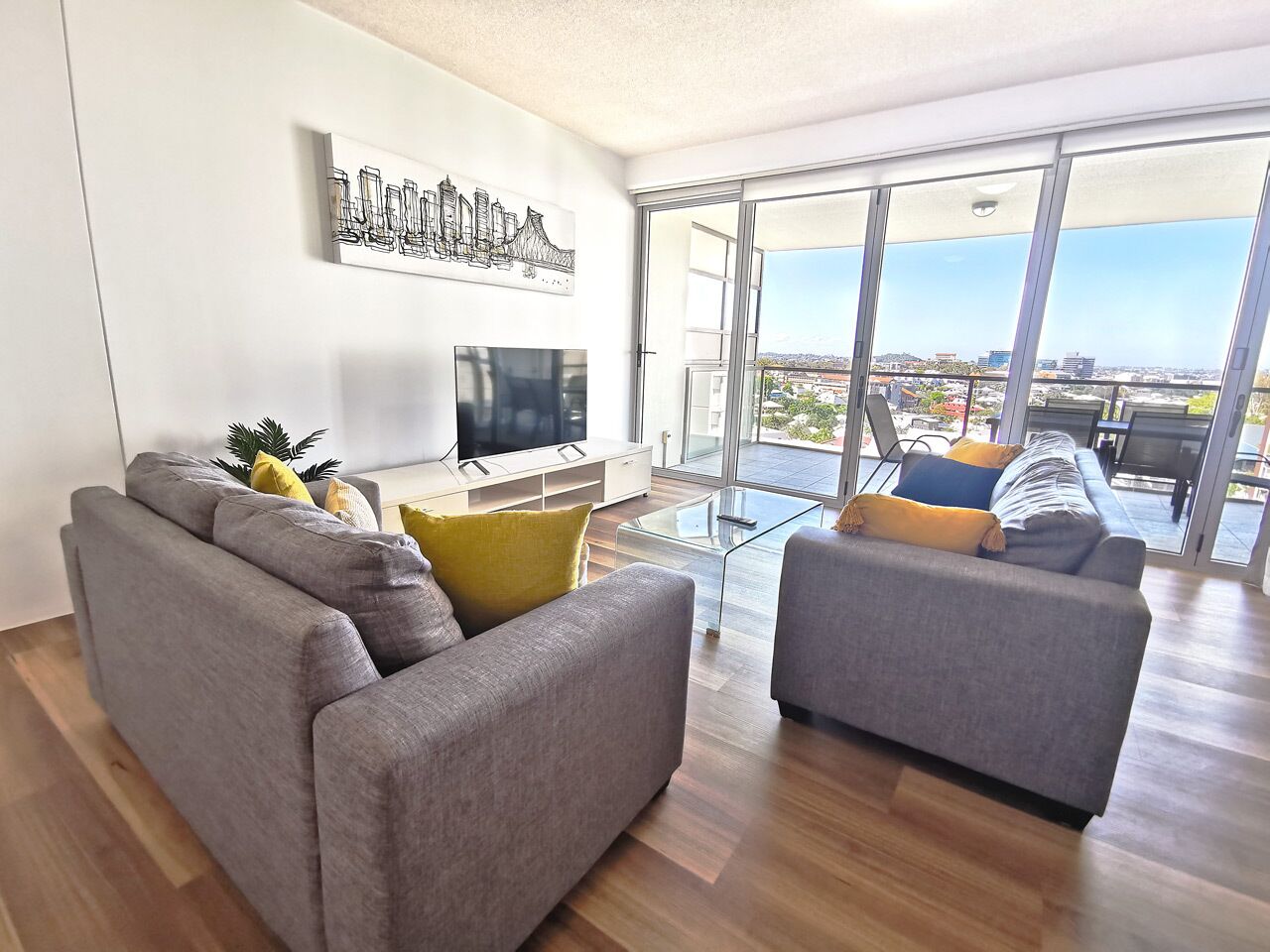 Una sala de estar moderna con un sofá gris, un sillón gris más pequeño y una mesa de centro de vidrio. Un televisor de pantalla plana está montado en la pared, y hay un ventanal con vistas a la ciudad.