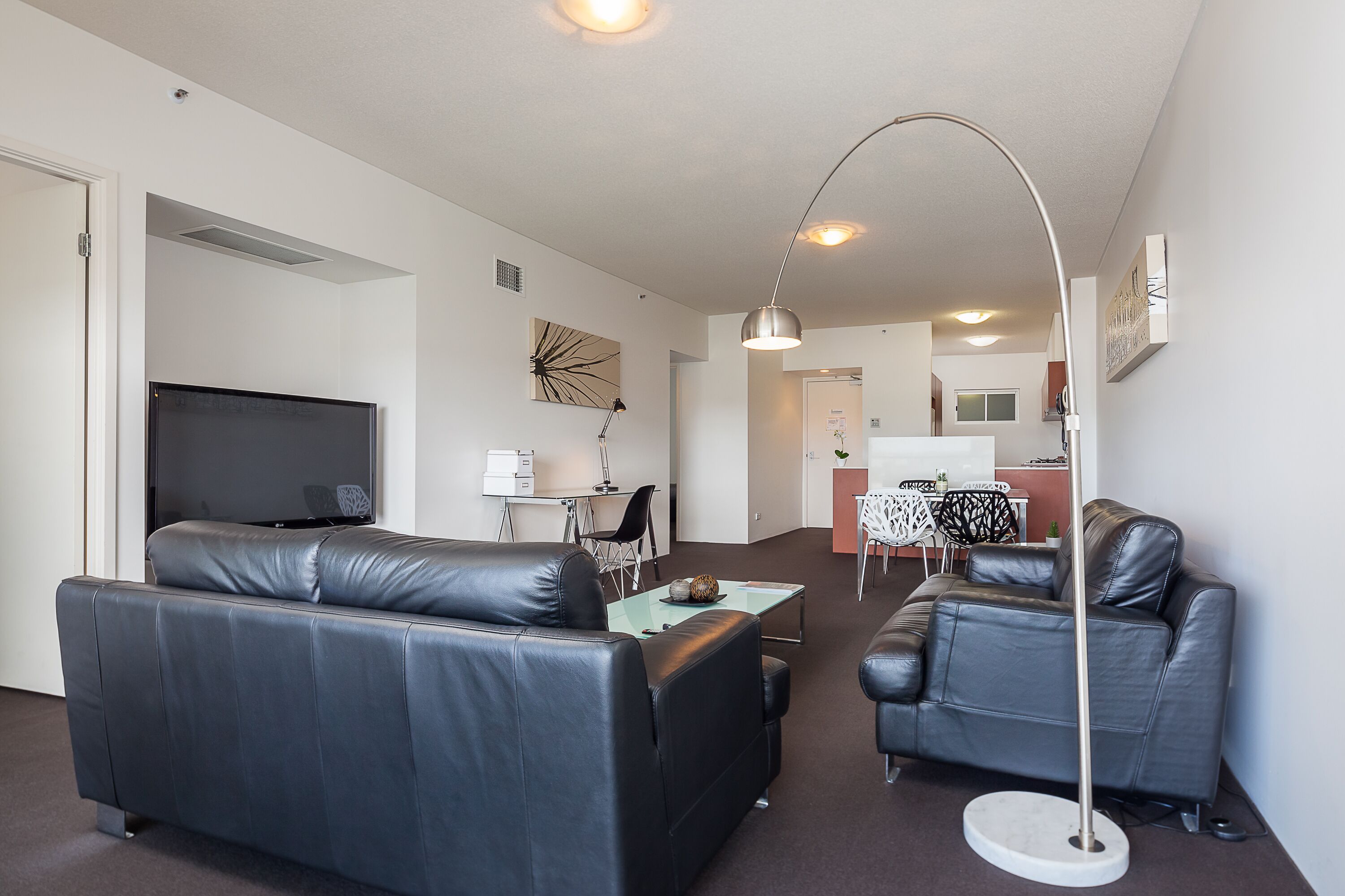 A modern living room with a large flat-screen TV, two leather sofas, a glass coffee table, and a floor lamp.