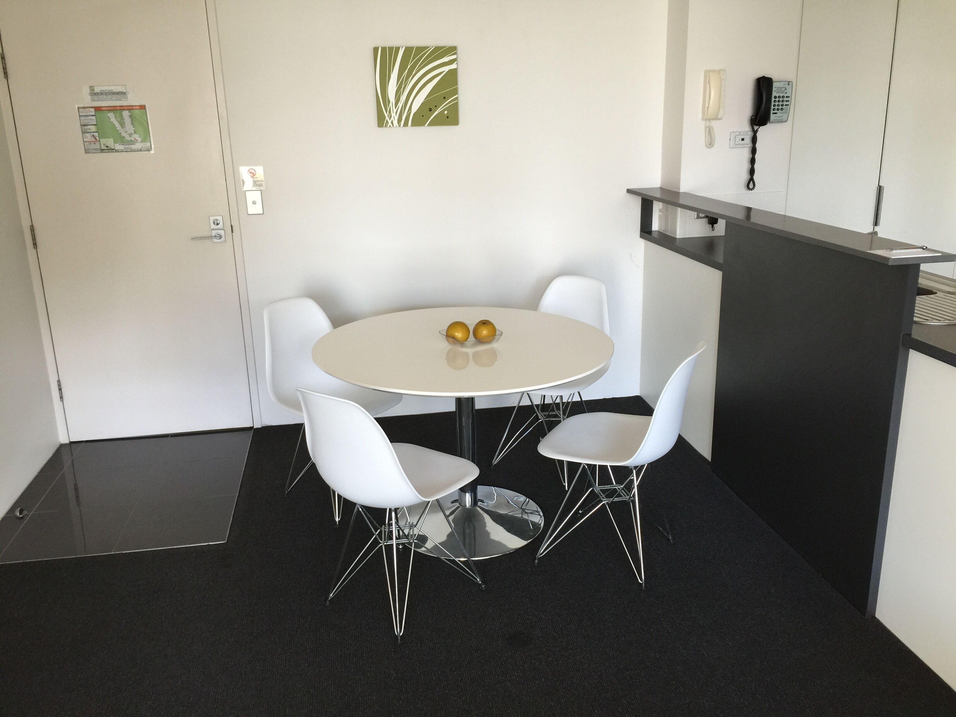 Un salón de conferencias con una mesa redonda, cuatro sillas blancas, dos manzanas sobre la mesa, un teléfono en la pared y una puerta con un cartel.
