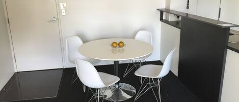 Un salón de conferencias con una mesa redonda, cuatro sillas blancas, dos manzanas sobre la mesa, un teléfono en la pared y una puerta con un cartel.