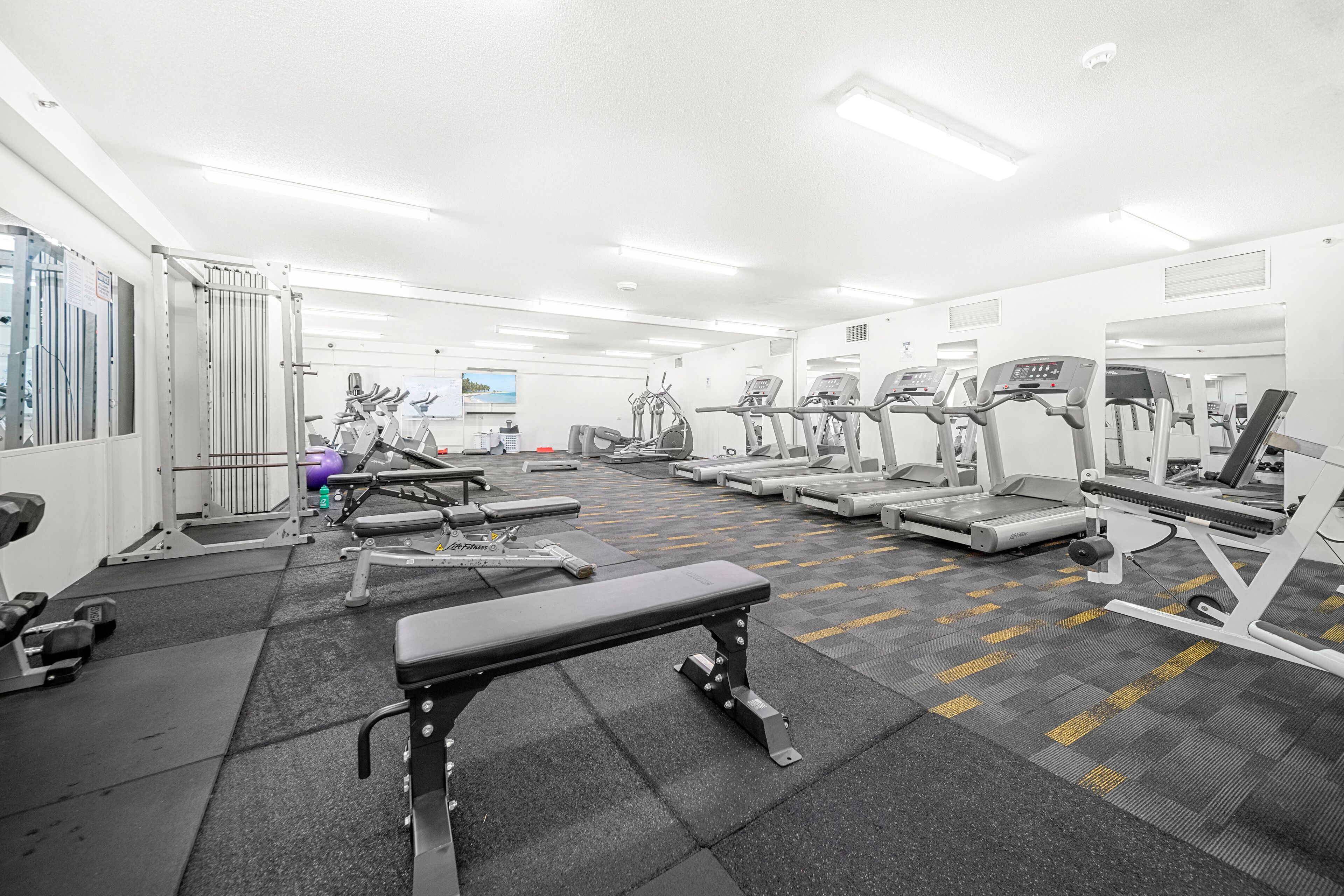 A gym with various exercise equipment, including treadmills, weight machines, and a bench.