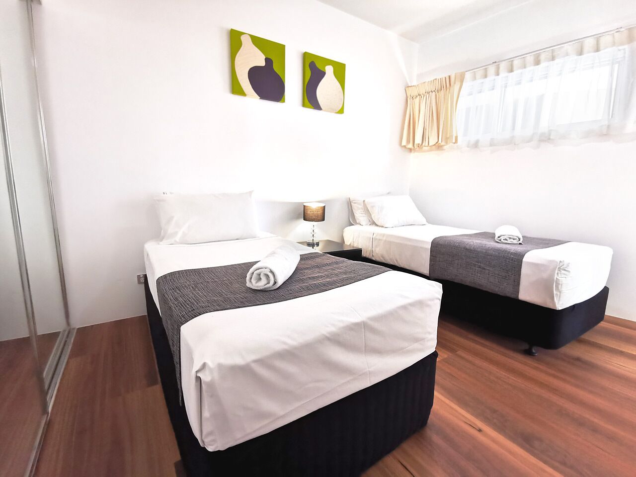 A hotel room with two beds, a bedside table with a lamp, and artwork on the wall.