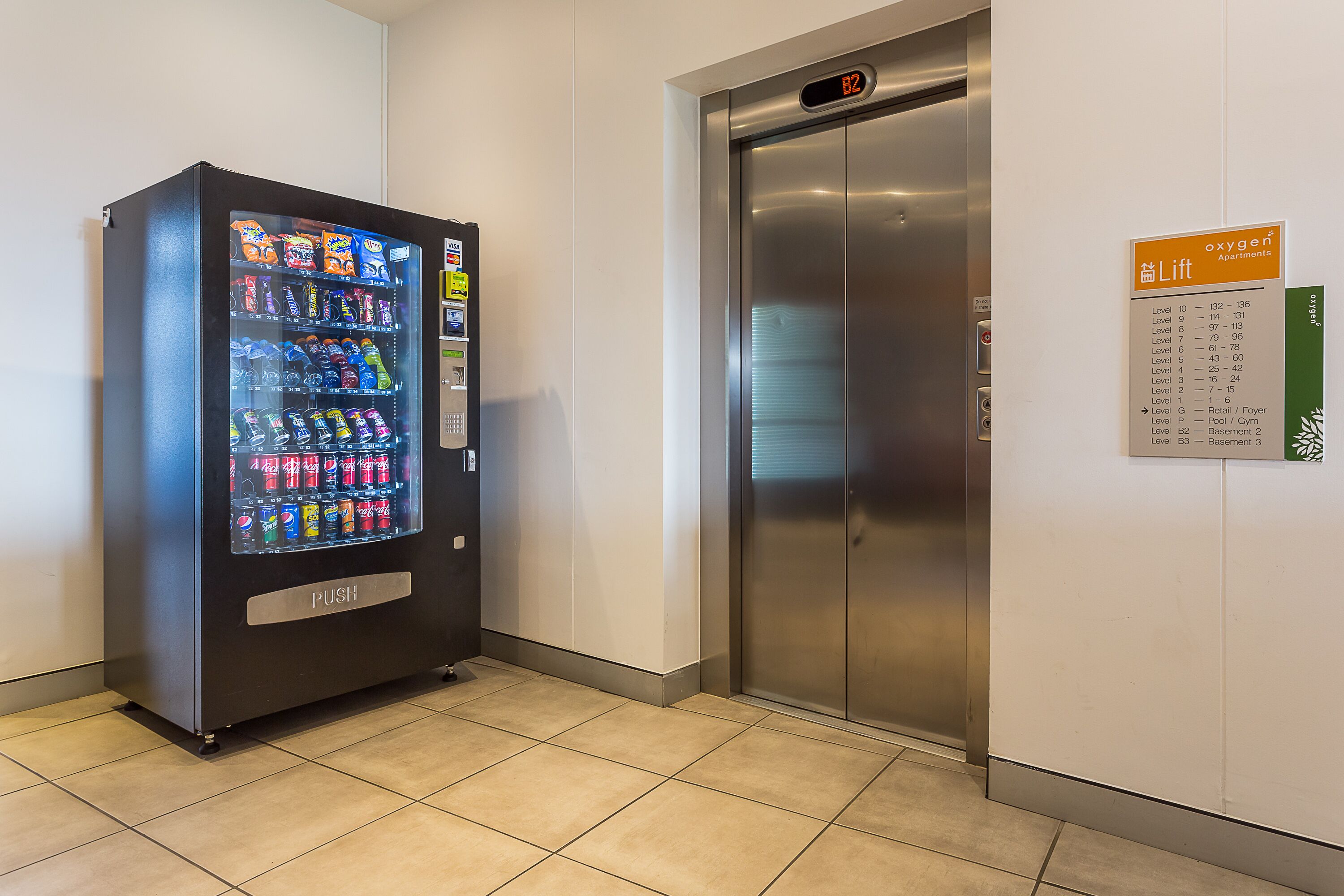 Una máquina expendedora con diversos aperitivos y bebidas, un ascensor con pantalla digital, y un cartel que indica el nombre y los pisos del edificio.