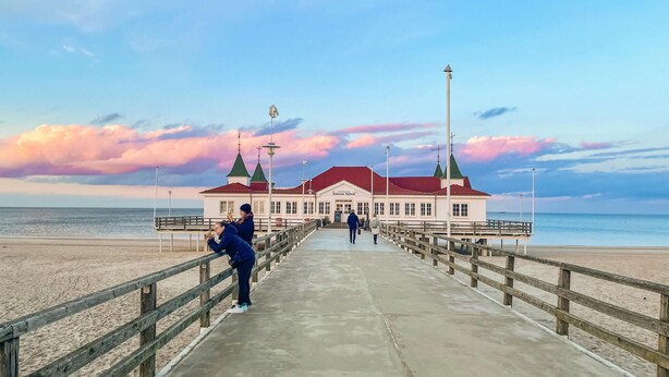 A pier leading to a building with a red roof and green towers, overlooking a beach.