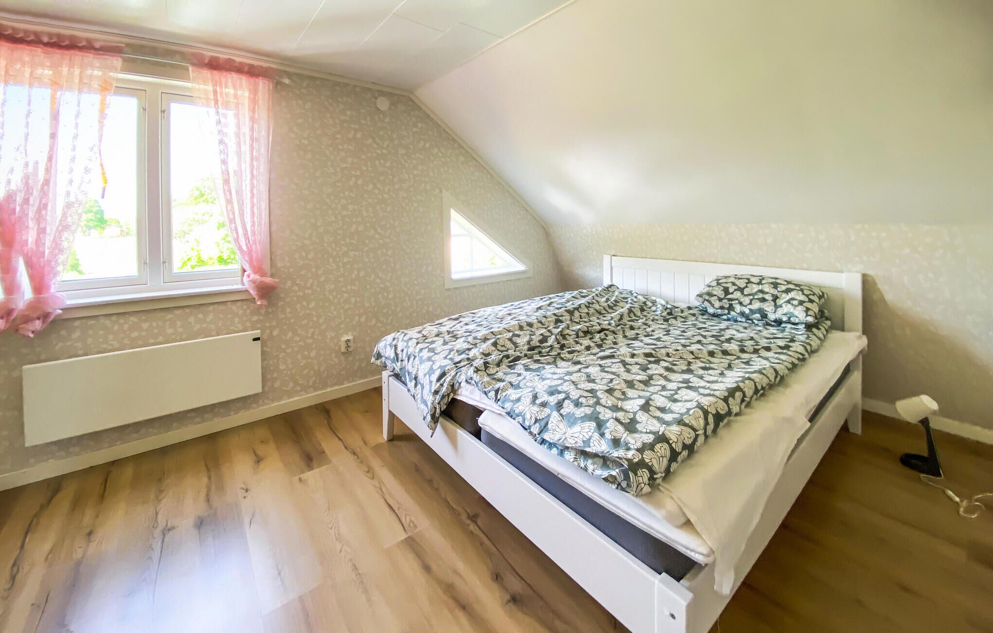A bedroom with a sloped ceiling, a bed with a floral bedspread, a radiator, and a window with pink curtains.