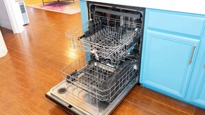 Uma máquina de lavar louça aberta com uma prateleira metálica interna, em uma cozinha com piso de madeira e armários azul claro.