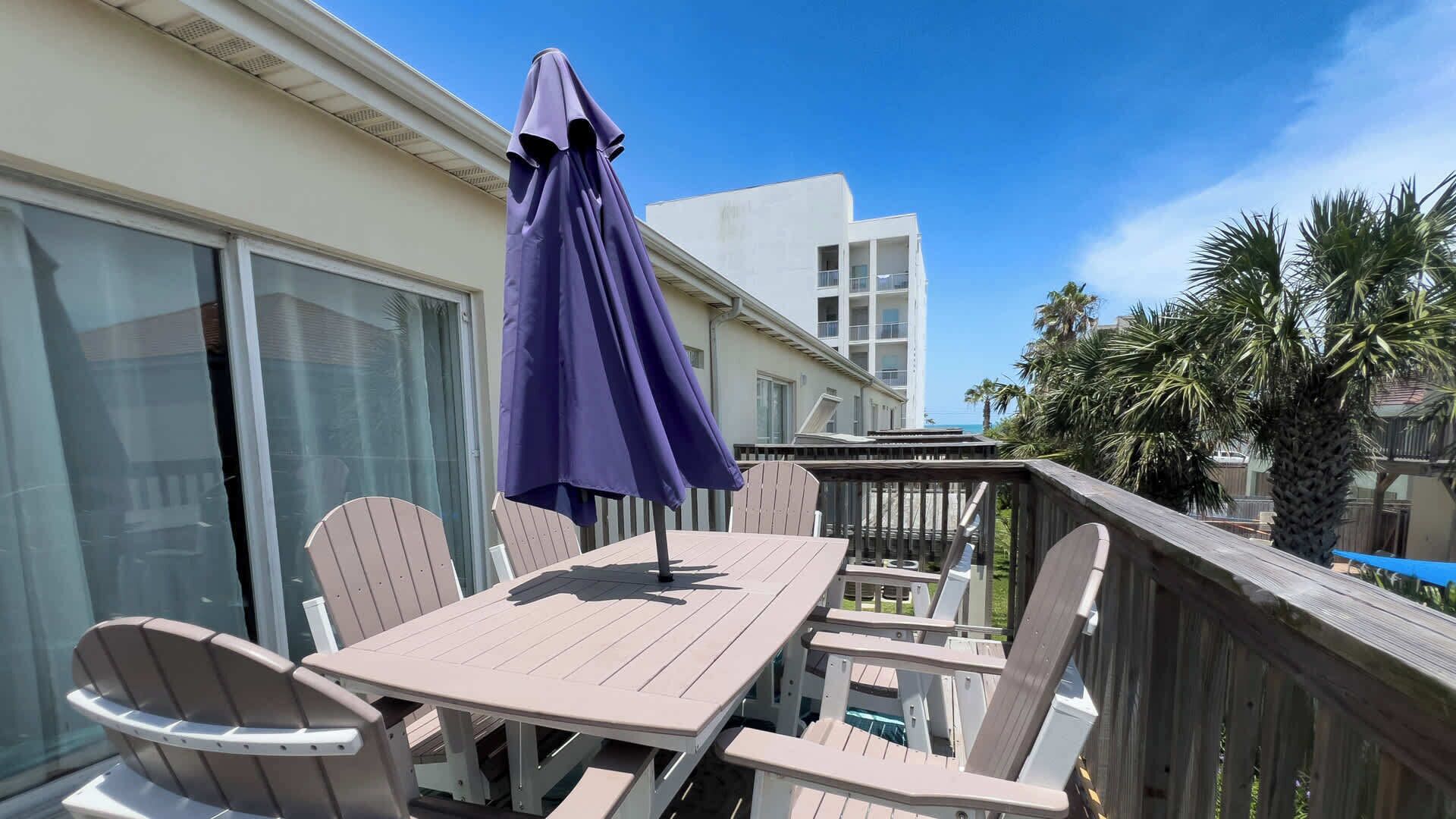 A balcony with a table and chairs, a purple umbrella, and a view of a building and palm trees.
