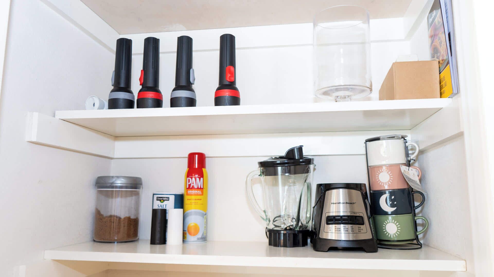 Ein Küchenregal mit verschiedenen Gegenständen, darunter Flaschen, ein Glas, ein Behälter, ein Mixer und eine Kaffeemaschine.