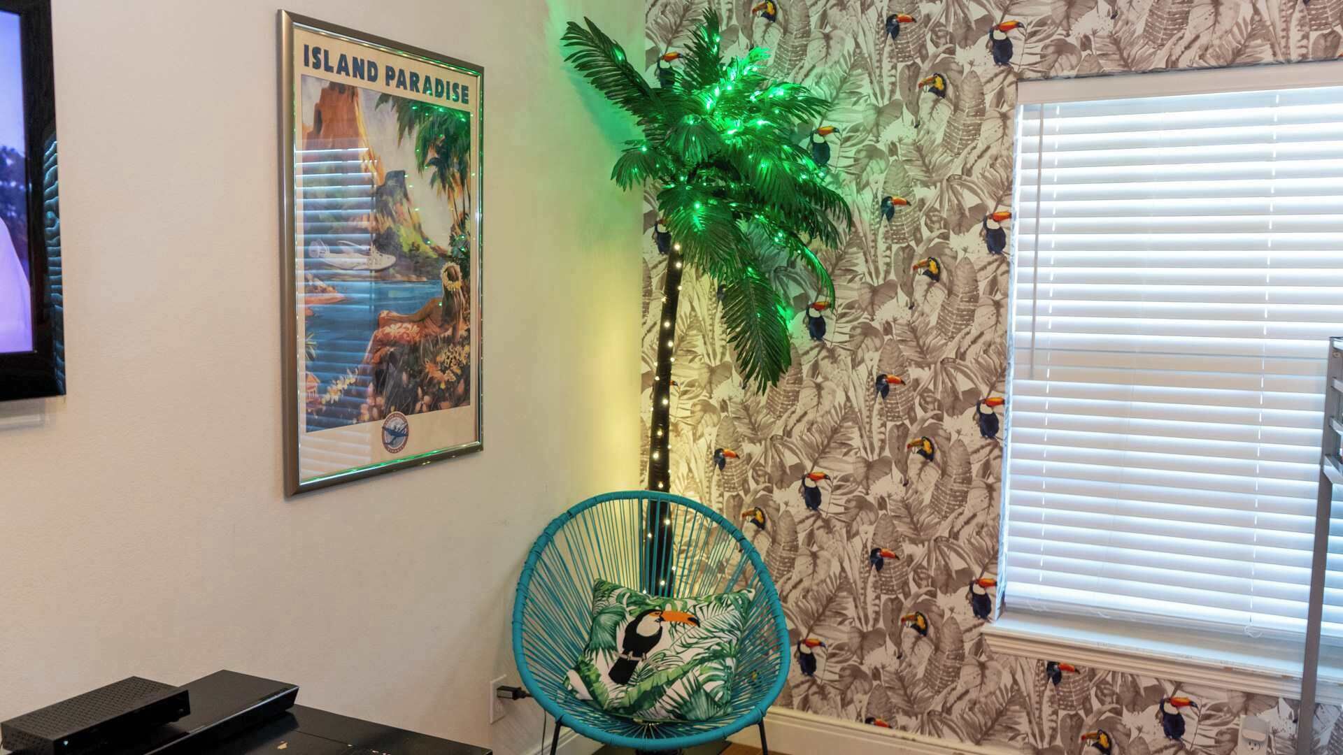 A room with a poster of 'Island Paradise', a green artificial palm tree, a chair with a tropical print cushion, and a patterned wallpaper with toucans.