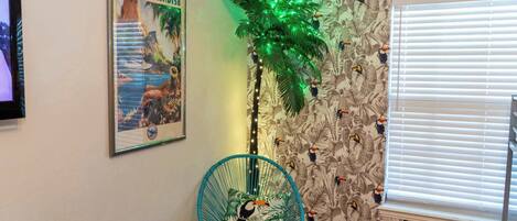 A room with a poster of 'Island Paradise', a green artificial palm tree, a chair with a tropical print cushion, and a patterned wallpaper with toucans.
