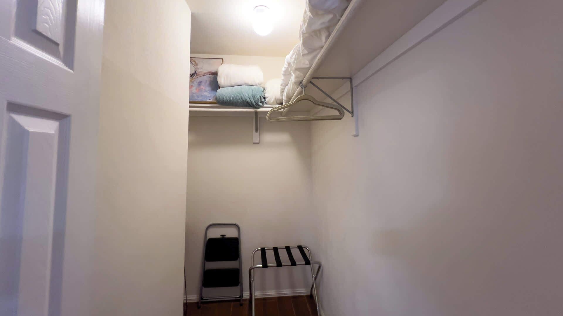 A narrow hallway with a white door on the left, a metal drying rack with a black towel, and a white shelf with a blue towel and a white pillow.