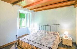 Ein Schlafzimmer mit Bett, einem Fenster mit grünen Lamellen, einem Nachttisch mit Lampe und einer Holzbalkendecke.