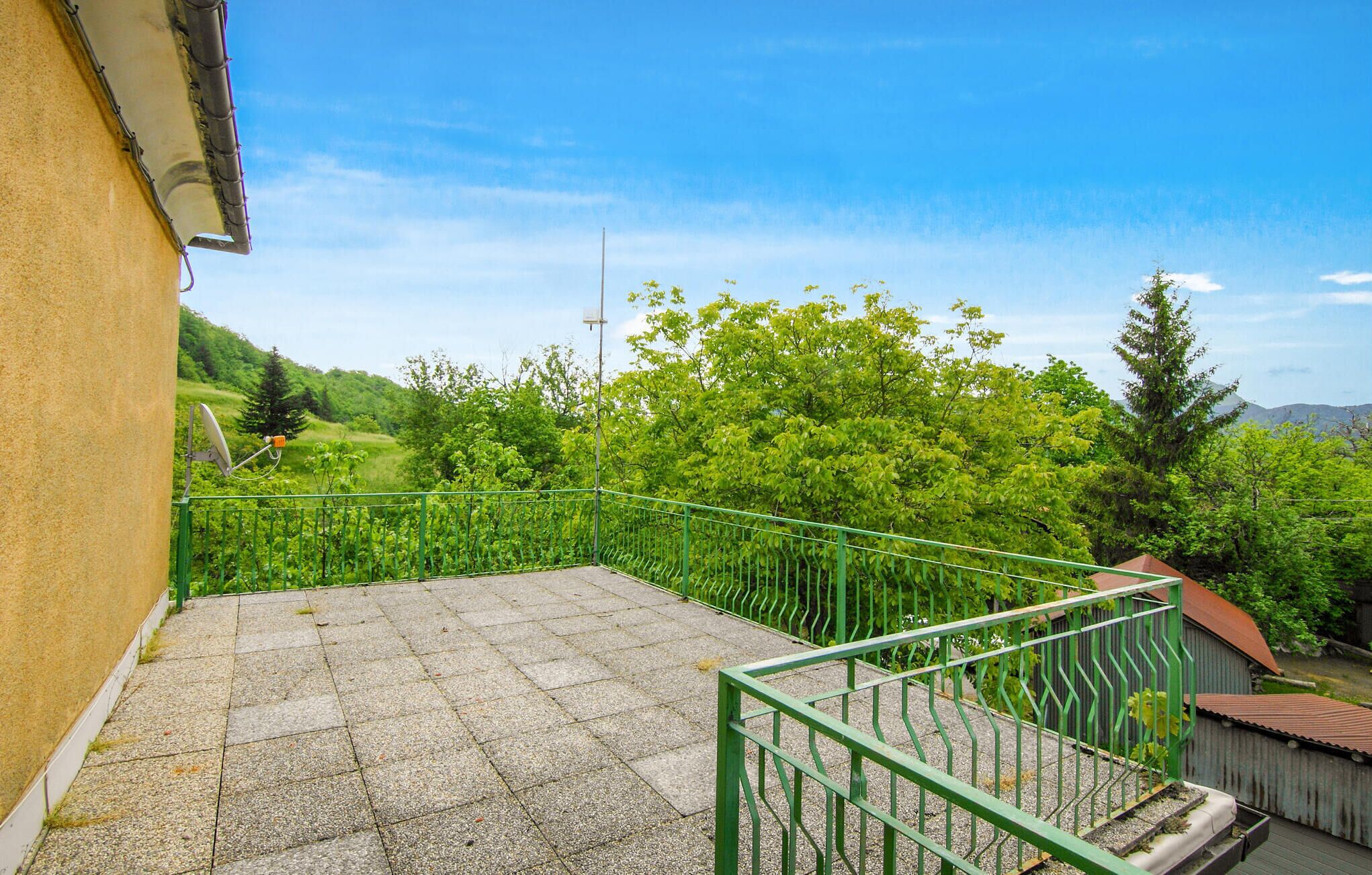 A balcony with a stone floor, green railing, and a view of a lush green landscape with trees and hills.