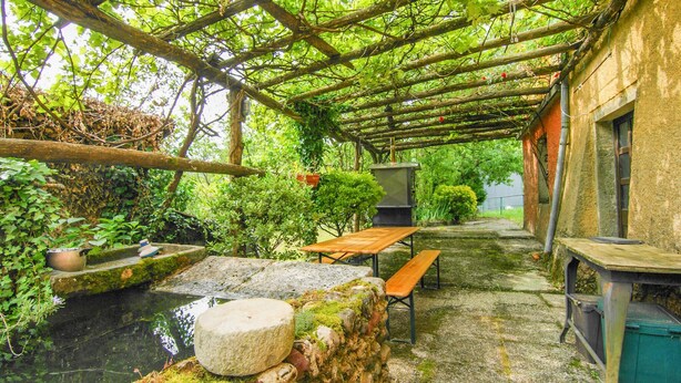 Un patio rústico al aire libre con una mesa y bancos de madera, rodeado de follaje y un muro de piedra.