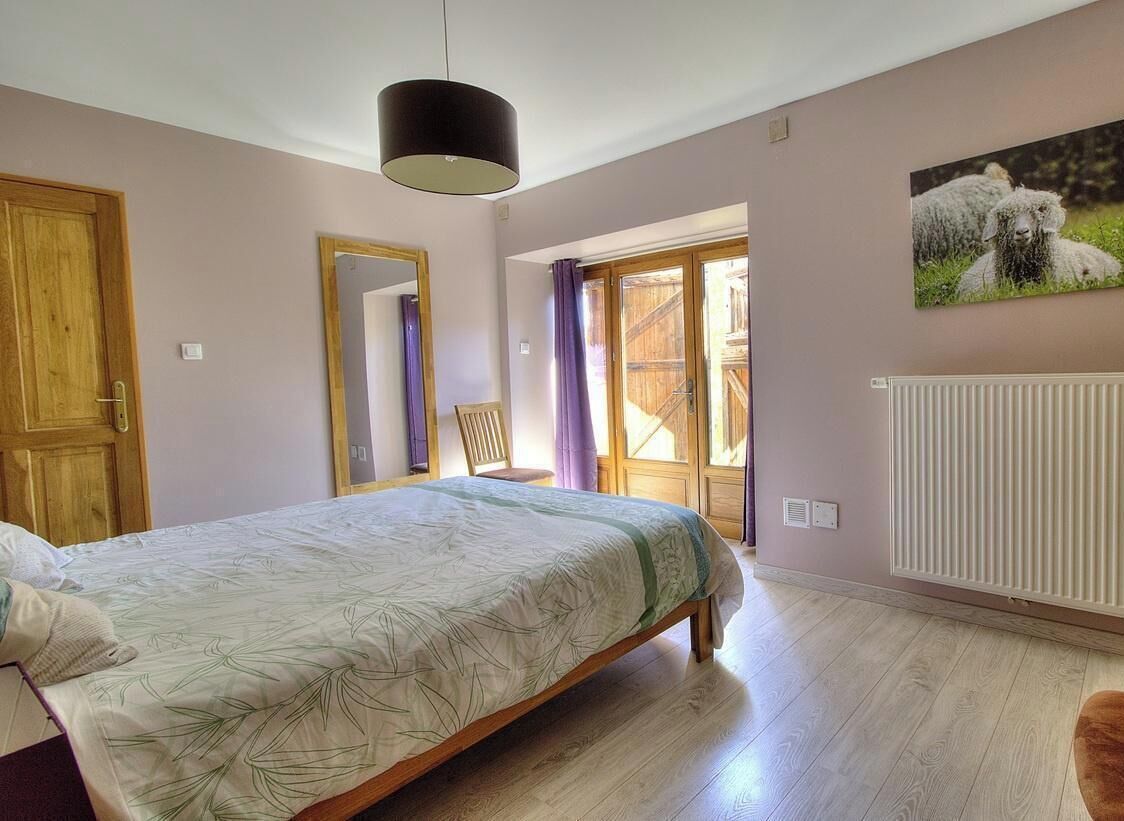Ein Schlafzimmer mit Bett, Spiegel, Fenster mit Vorhängen, Heizkörper und einem Bild an der Wand.