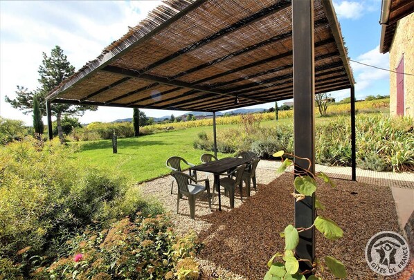 A covered outdoor seating area with a table and chairs, surrounded by a garden with various plants and a view of a vineyard in the background.