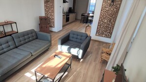 Ein modernes Wohnzimmer mit einem grauen Sofa, einem kleineren grauen Sessel und einem Holztisch. Im Hintergrund ist ein Essbereich mit Tisch und Stühlen erkennbar.