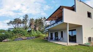 Une maison moderne à deux étages, dotée d’un balcon couvert, d’une pelouse et entourée d’arbres.
