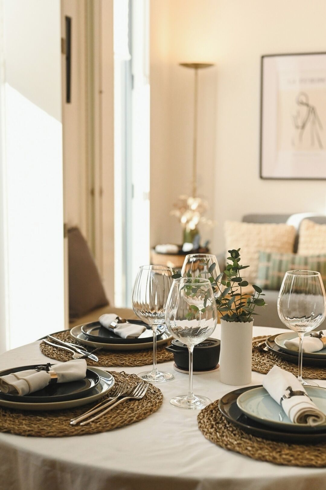 A dining table set with plates, cutlery, and wine glasses, with a vase of greenery and a framed picture on the wall.