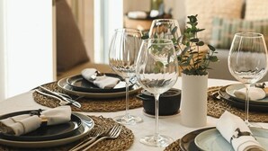 Una mesa de comedor con platos, cubiertos y copas de vino, con un florero con follaje y un cuadro en la pared.
