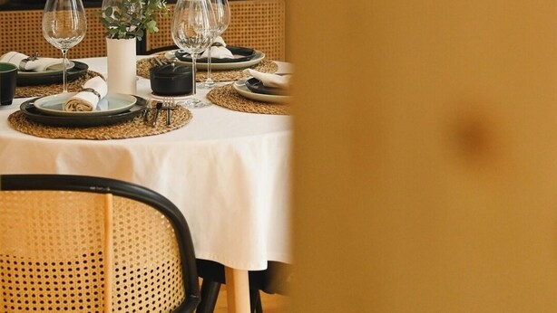 Un comedor con una mesa dispuesta para cuatro, incluyendo copas de vino, platos y cubiertos. Hay individuales tejidos, un florero con follaje y una vela sobre la mesa. La habitación tiene una paleta de colores cálidos, con una silla de mimbre y un cuadro con marco negro sobre la mesa.