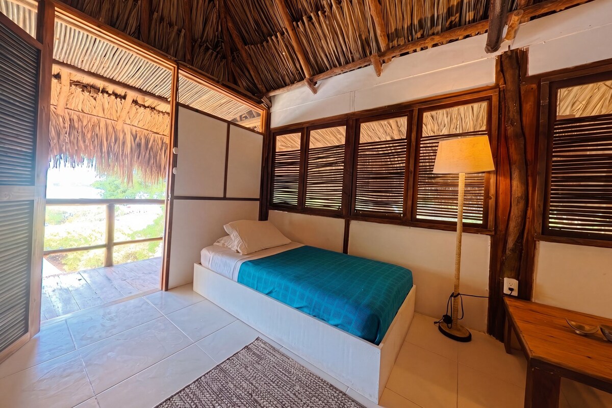 A room with a bed, a window with blinds, a lamp, and a view of greenery outside.