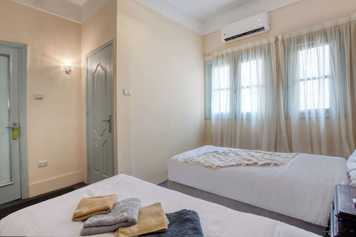 A hotel room with two beds, a wardrobe, an air conditioning unit, and a window with curtains.