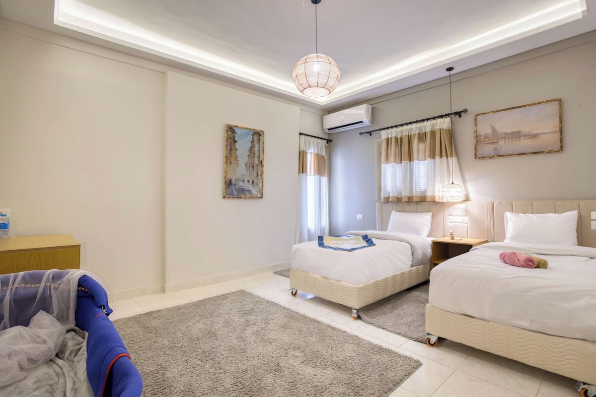 A hotel room with two beds, a large window with curtains, a ceiling light, an air conditioning unit, and a painting on the wall.