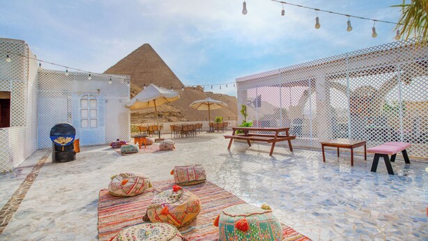 An outdoor seating area with patterned cushions, a wooden bench, and a view of a pyramid. - Villa Khufu Pyramids Hotel (Giza)
