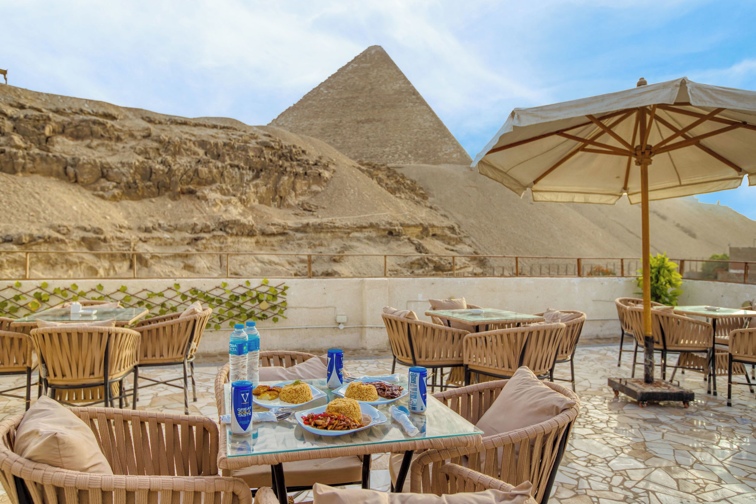 Un'area pranzo all'aperto con sedie e tavoli in vimini, un ombrellone e vista sulla Grande Piramide di Giza sullo sfondo.