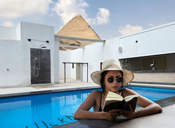 A person reading by a pool with a modern building and a pyramid in the background.