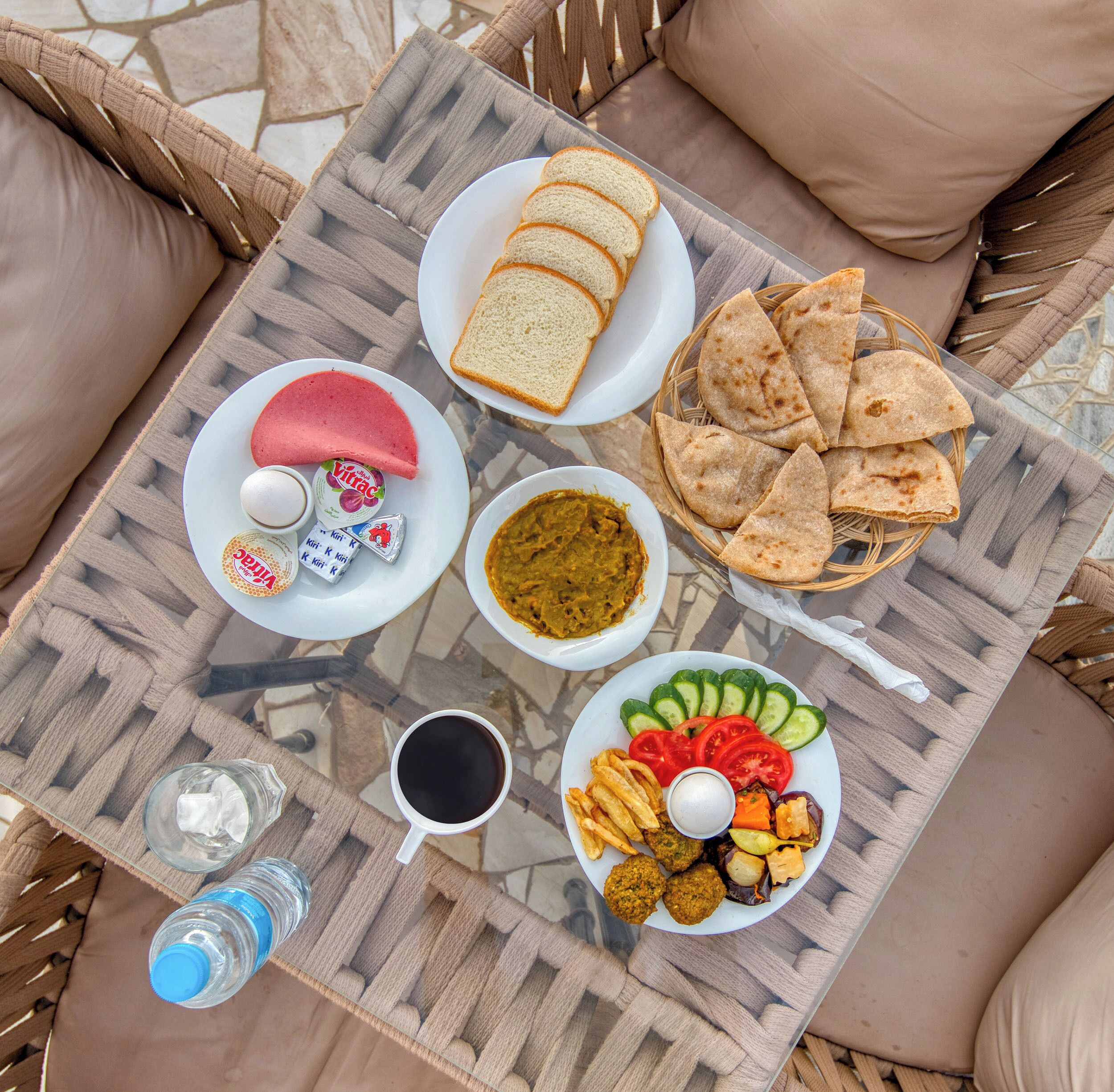A table with various food items including bread, flatbreads, a plate with vegetables, a plate with a red dessert, a plate with a green dessert, a plate with fried items, a cup of coffee, and a bottle of water.
